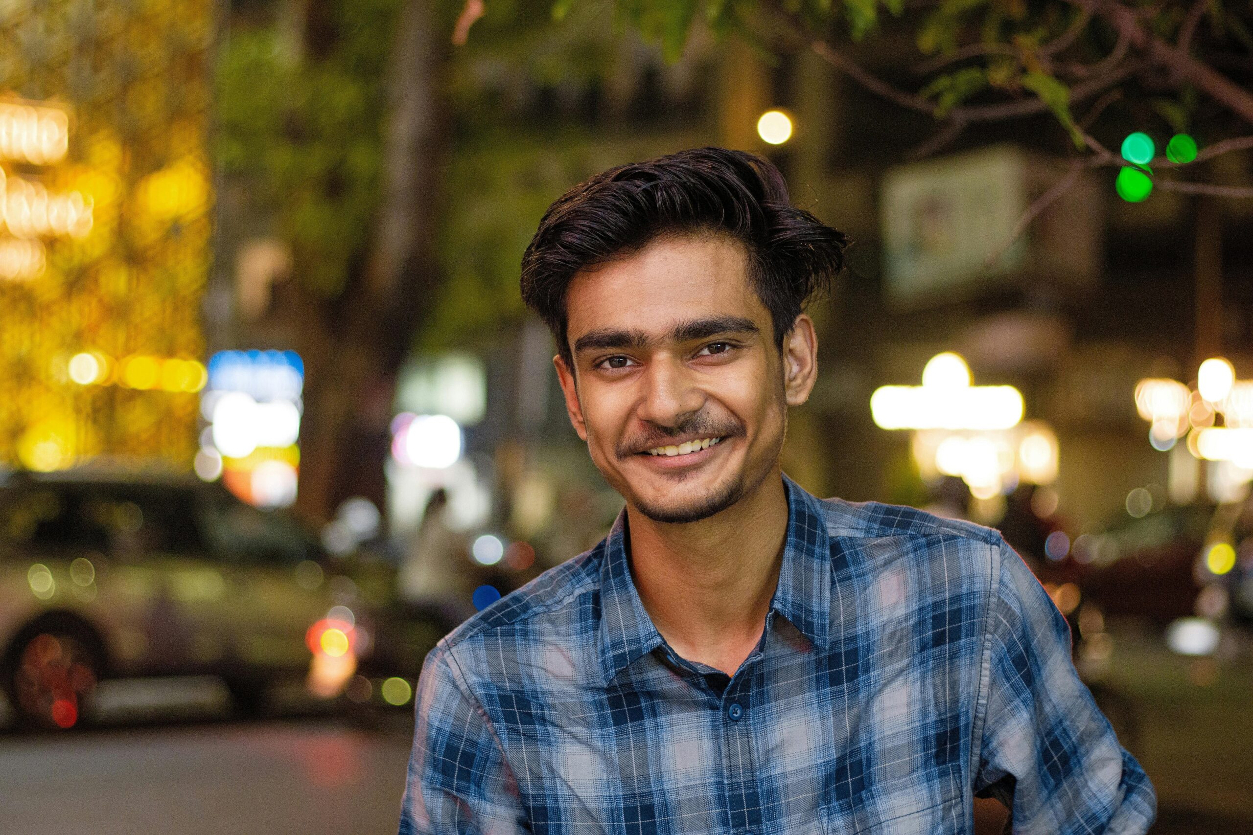A cheerful young man in a plaid shirt smiles at the camera in an urban night scene.